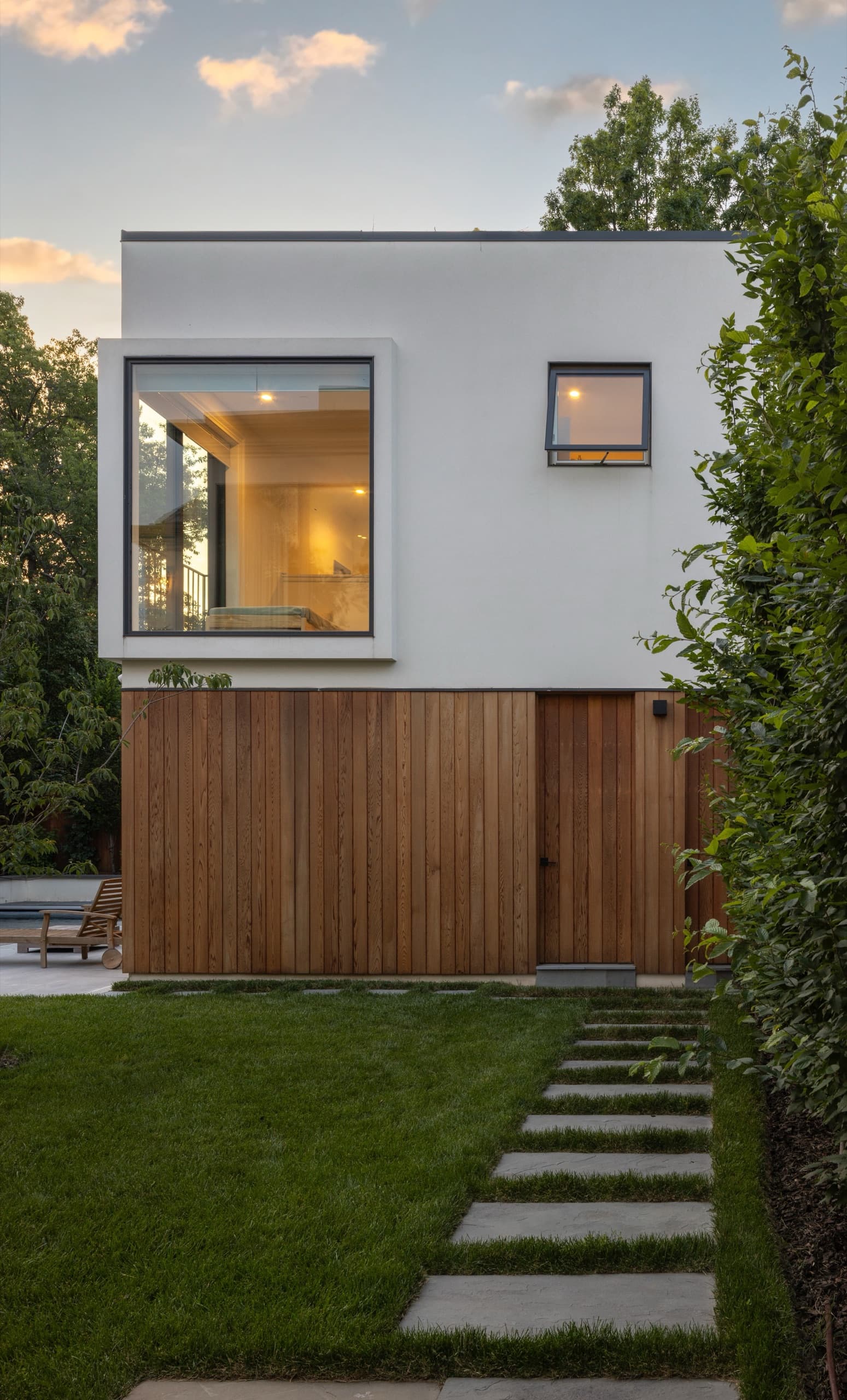 Timber garage door set into a rendered white volume, with stone stepping stones across the lawn